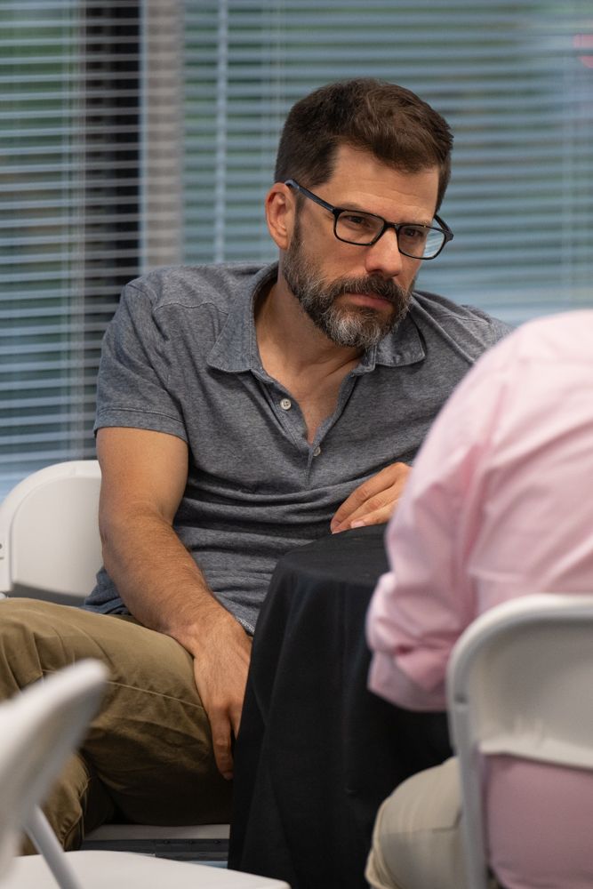 Professional wearing glasses and grey shirt leans forward during business meeting discussion at office desk.