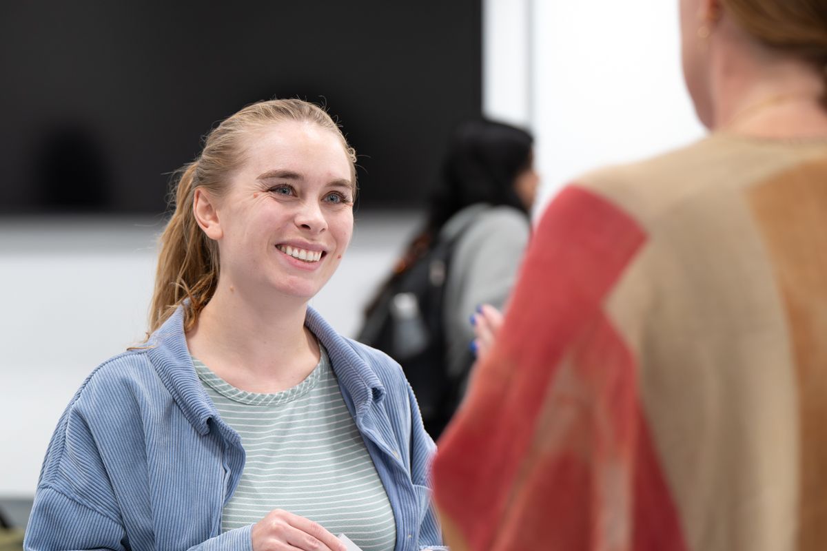 Person in denim jacket and striped shirt smiles during conversation in bright indoor setting.