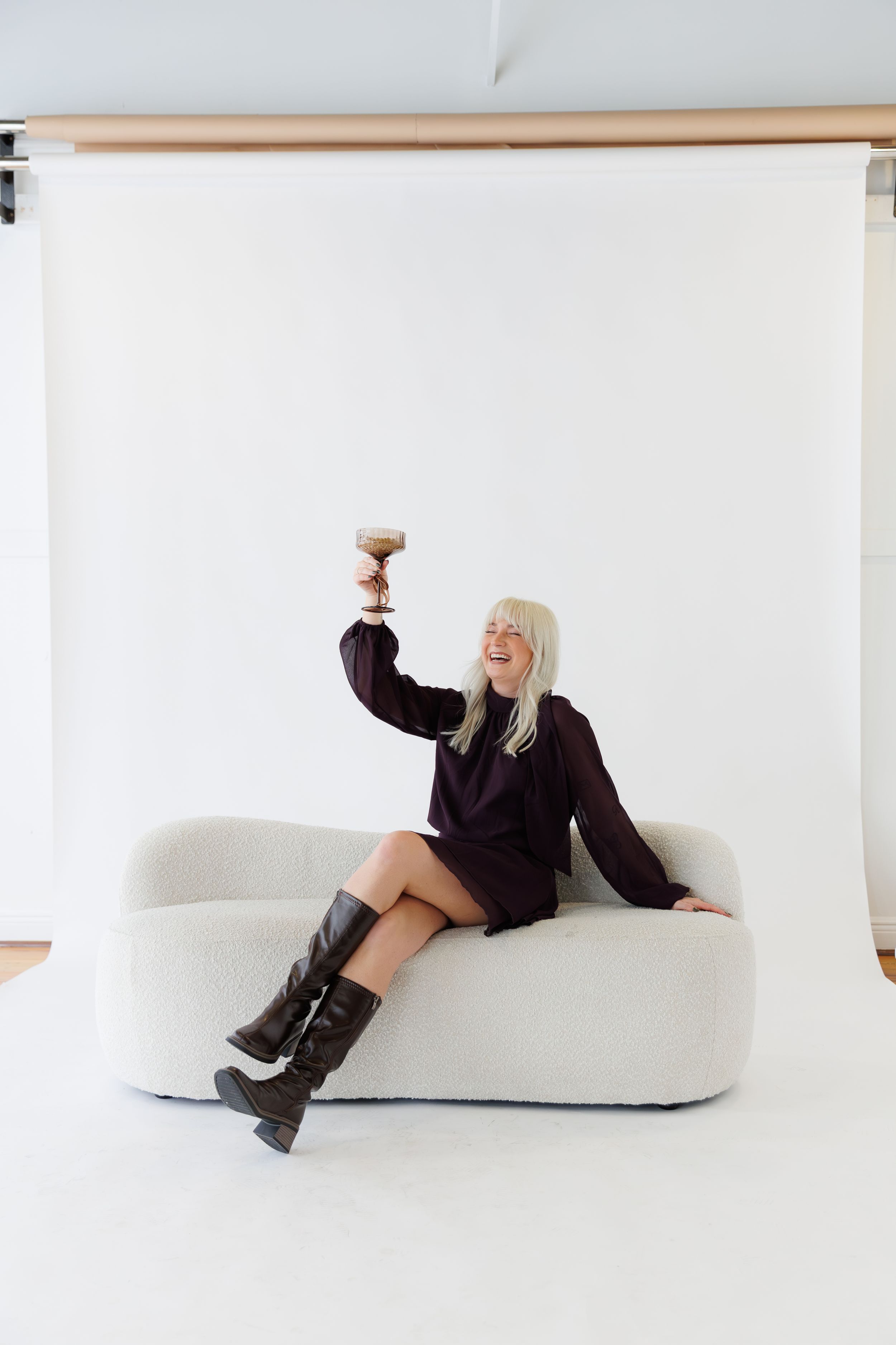 Person in black outfit lounging playfully on white modern furniture against minimalist white backdrop.