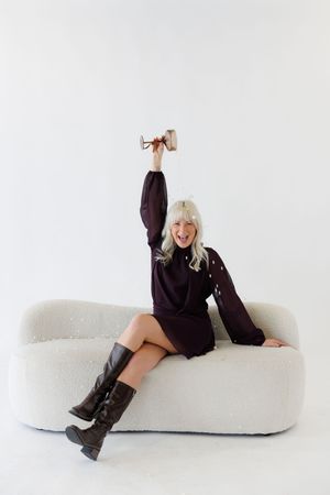 A person in a black sweater poses playfully on a white modern chaise lounge in a minimalist setting.