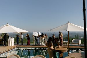 People gather under white tents and umbrellas at an outdoor event with mountain views in the background.