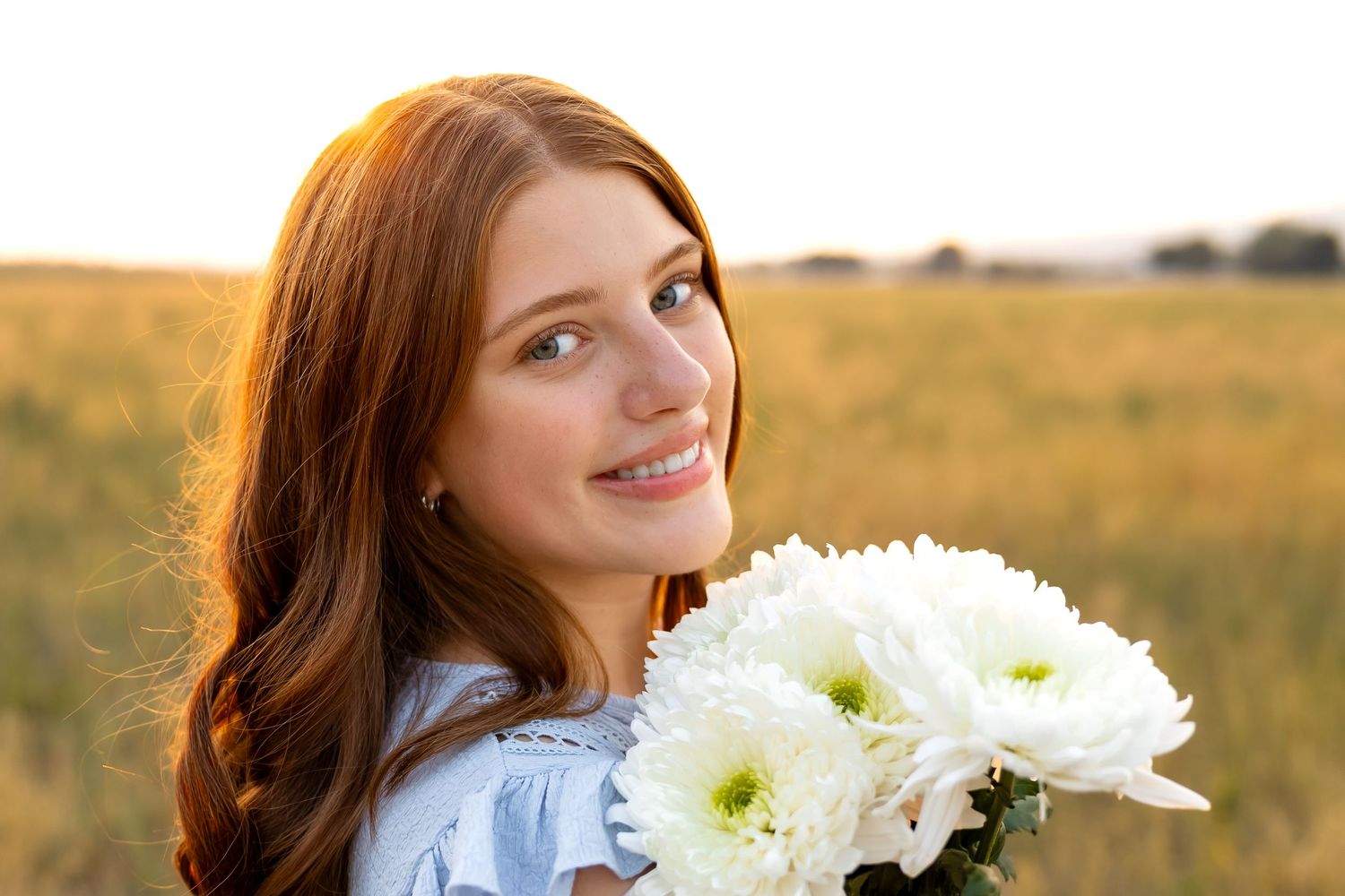 Woman with long auburn hair poses in a sunny field while holding white chrysanthemum flowers.