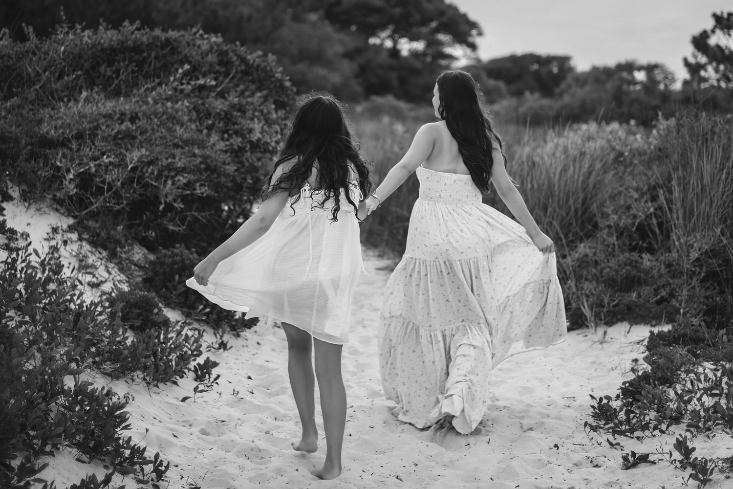 Two women in white dresses walking hand in hand through sandy beach dunes in black and white.