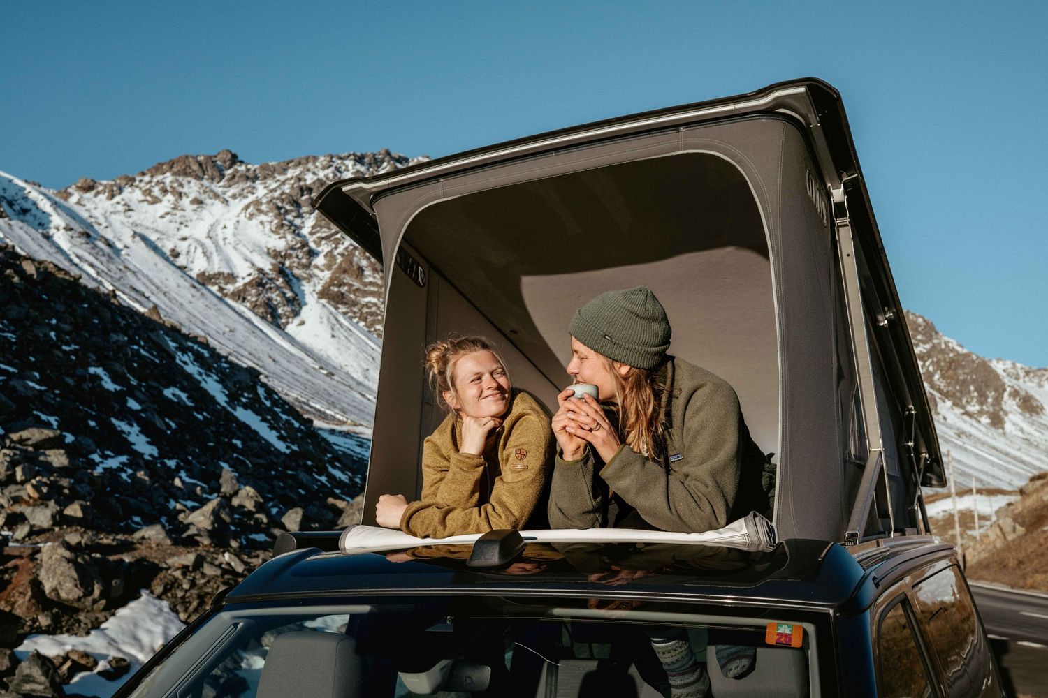 Two people enjoy coffee and snacks from their rooftop tent while winter camping in snowy mountain terrain.