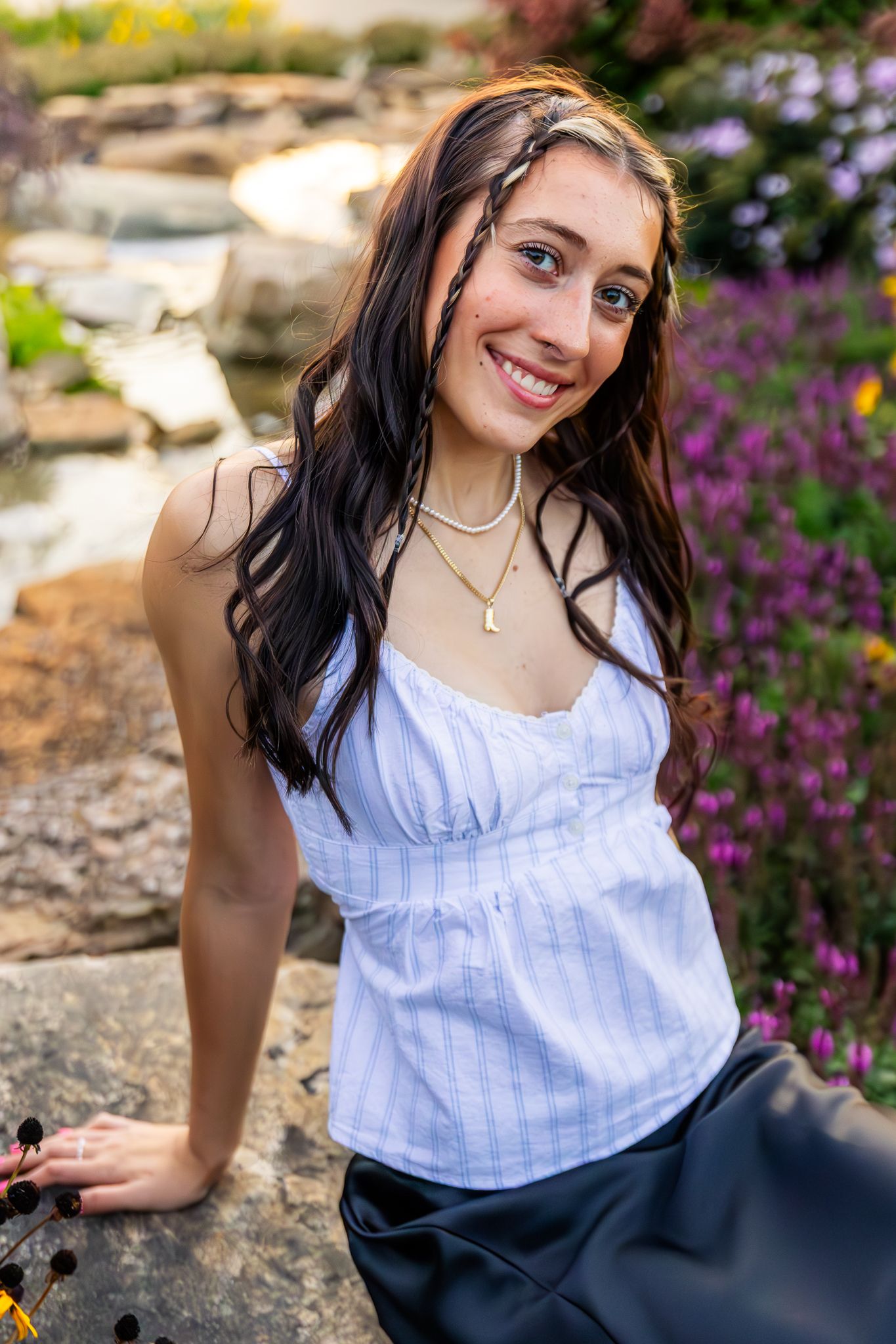 Senior girl sits for a senior photo at golden hour near a stream. She is sitting in front of flowers at Four Star Greenhouse.