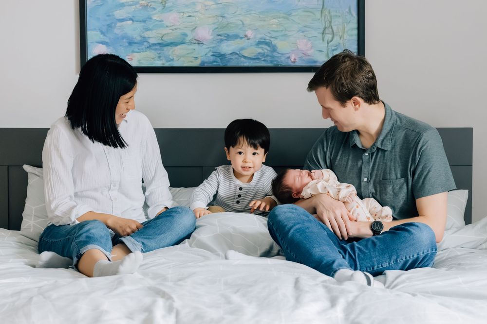 Family of four sitting on bed with toddler boy and newborn baby girl in cozy bedroom.