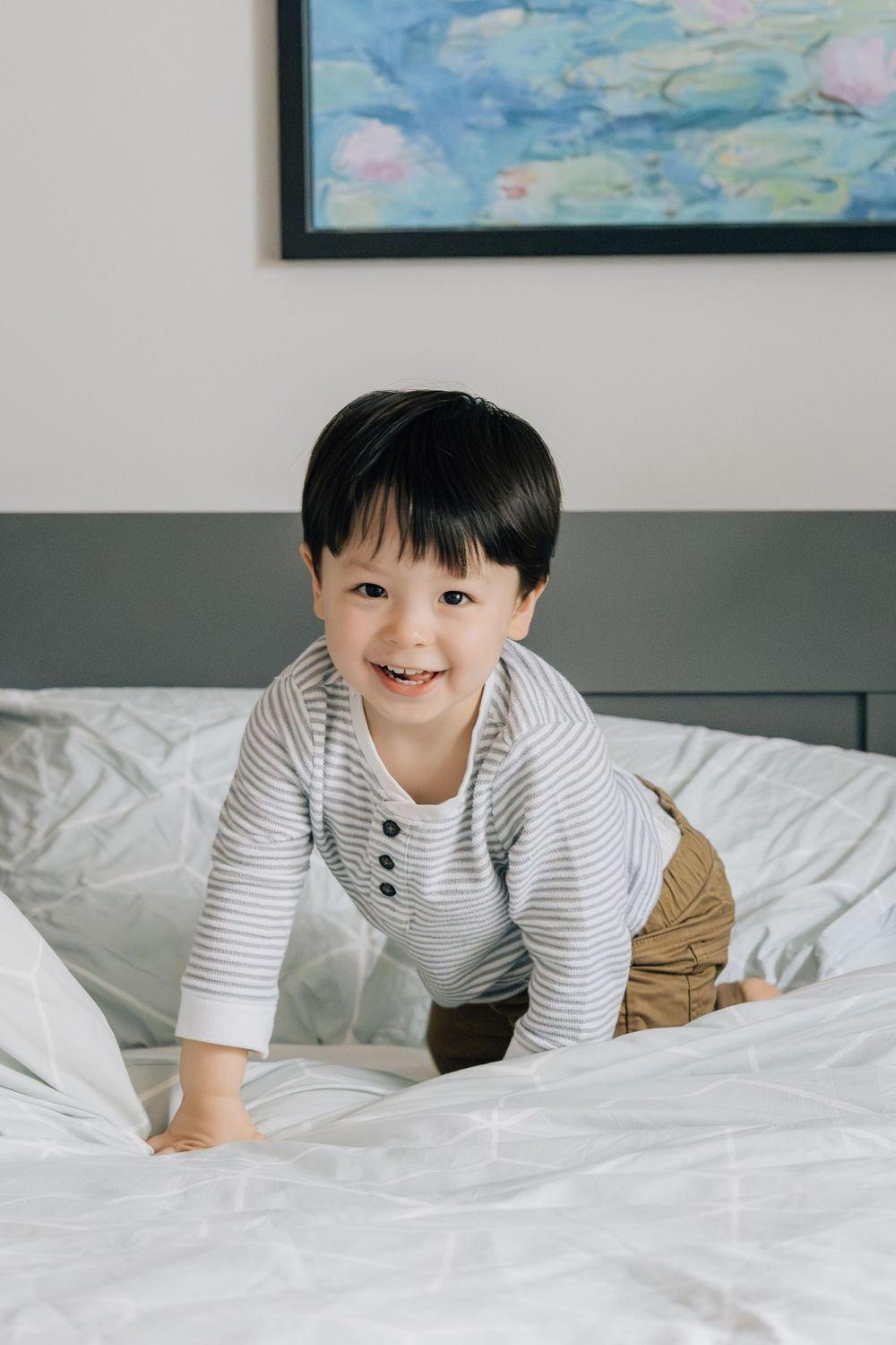 Adorable smiling Asian toddler boy in striped shirt crawling on white bed with artwork behind.