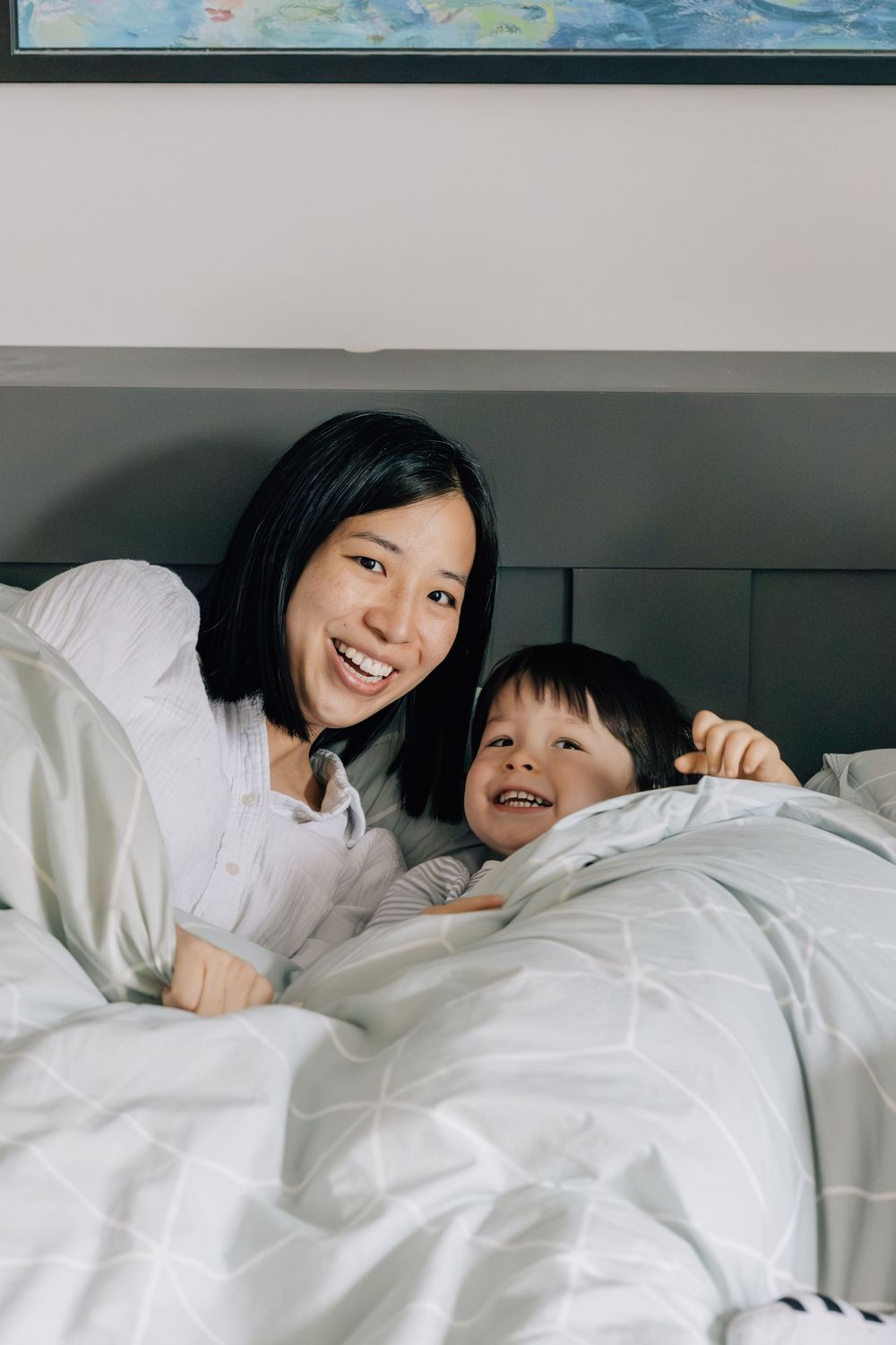 Smiling Asian mother and young toddler son snuggled together under white bedding covers.