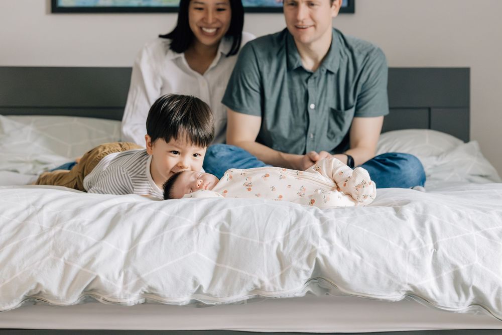 Parents watching lovingly as toddler son lies next to newborn baby sister on white bed.
