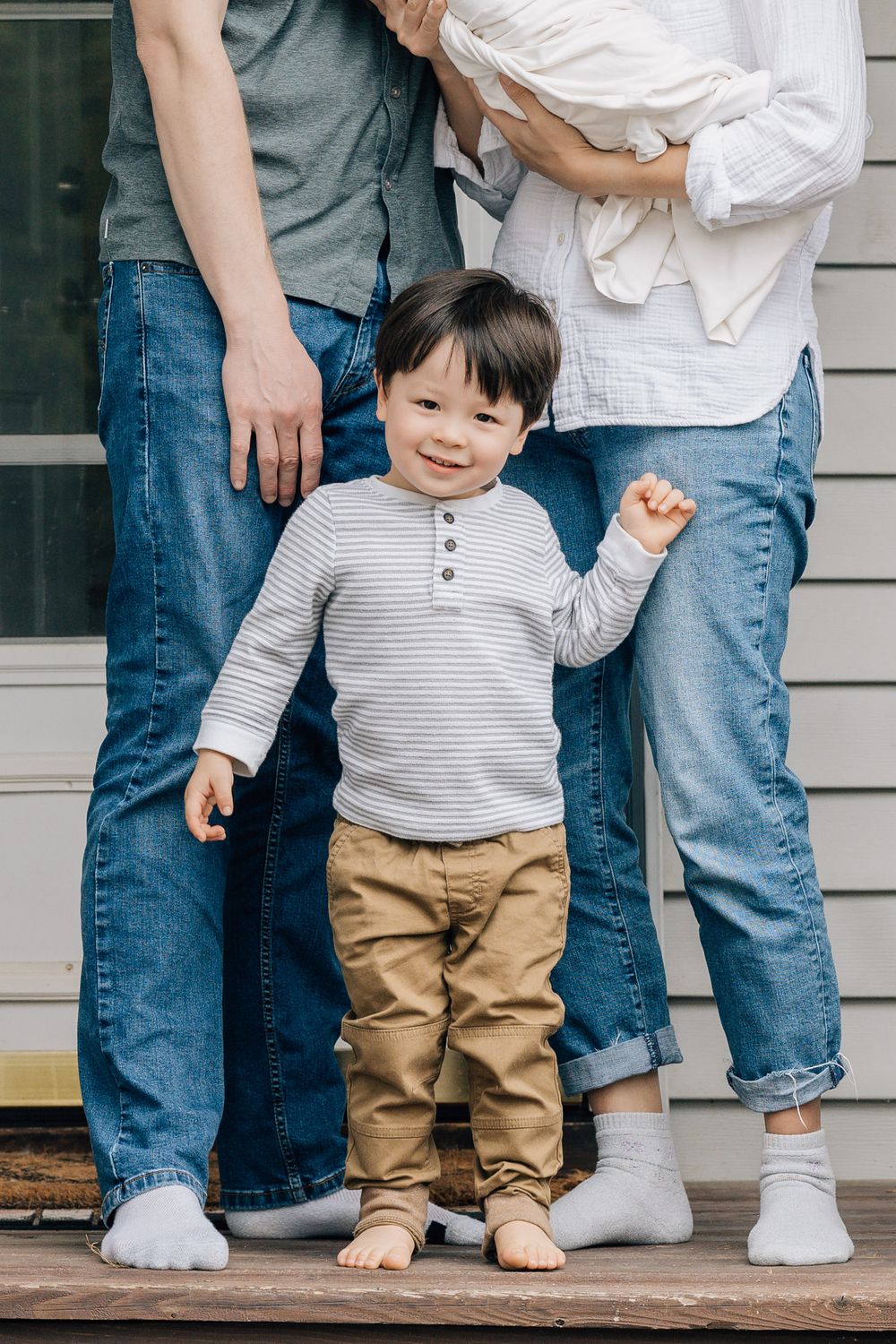 Smiling toddler boy in striped shirt standing between parents legs on wooden porch steps.