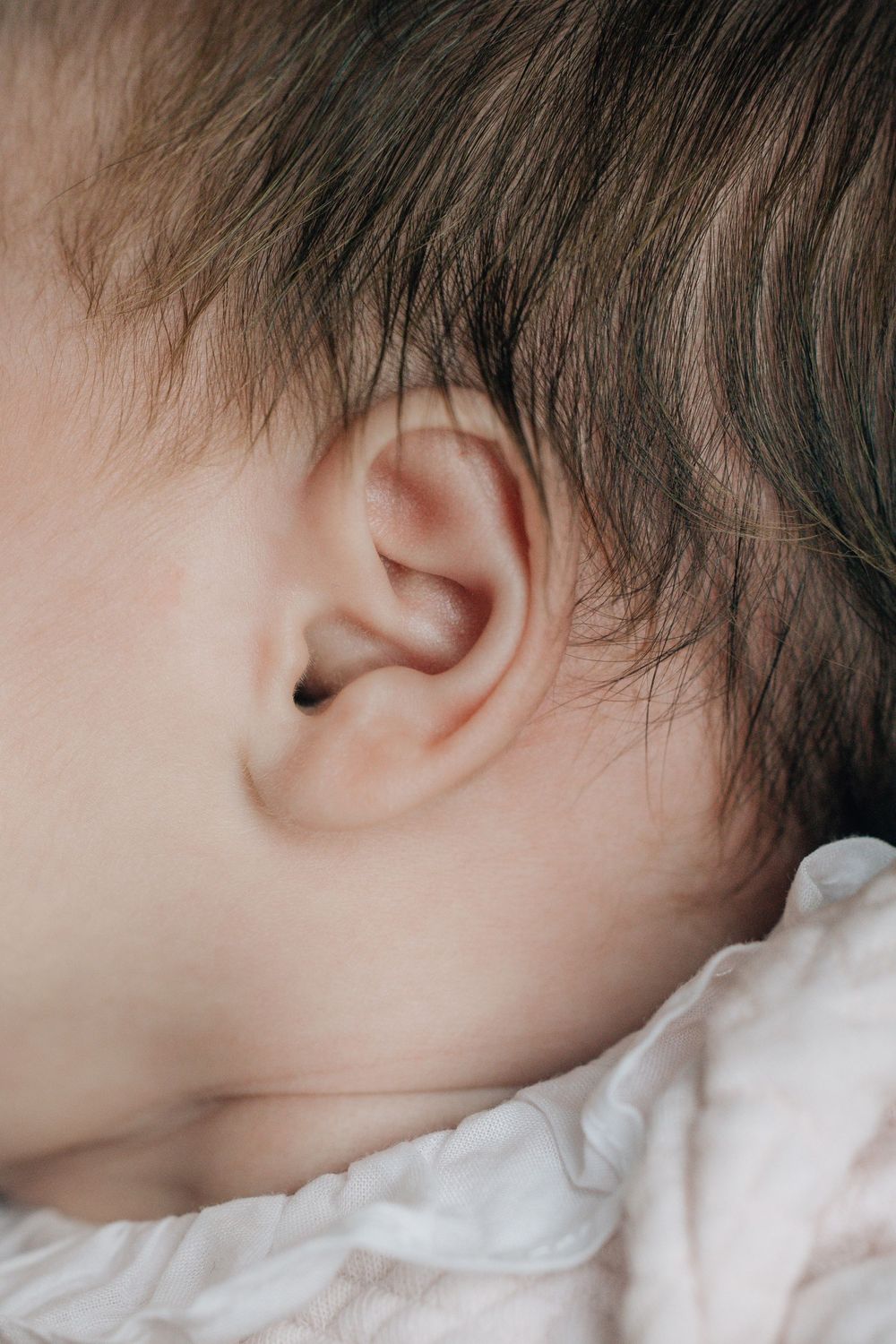 Close-up detail of sleeping newborn baby ear with soft dark hair on white blanket.