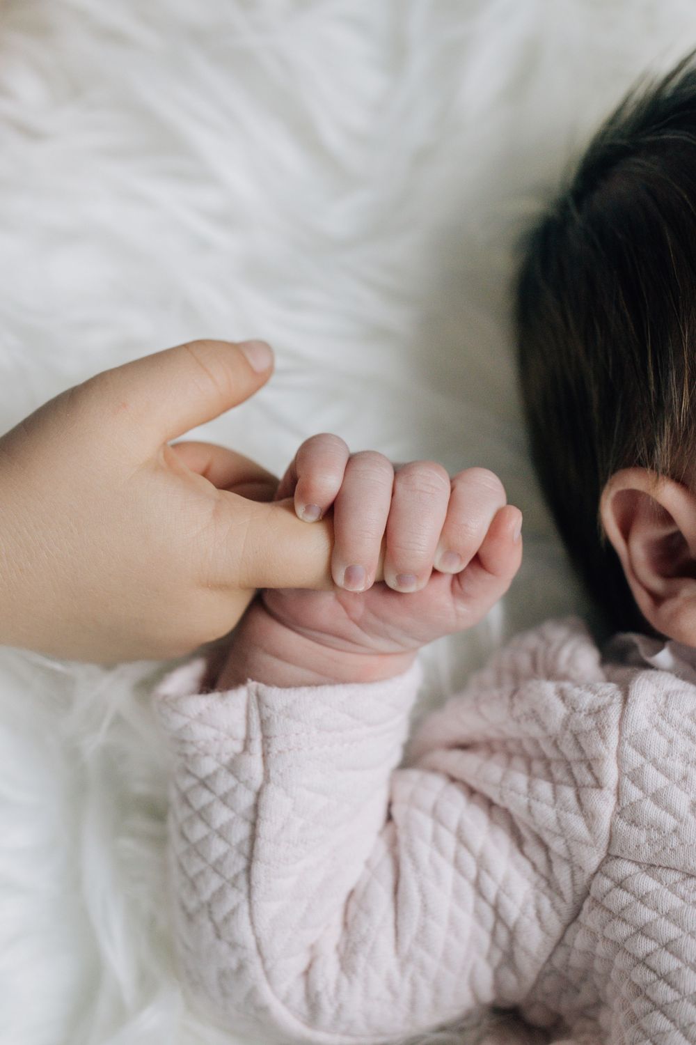 Newborn baby in pink outfit grasping toddler sibling finger on soft white fur blanket.