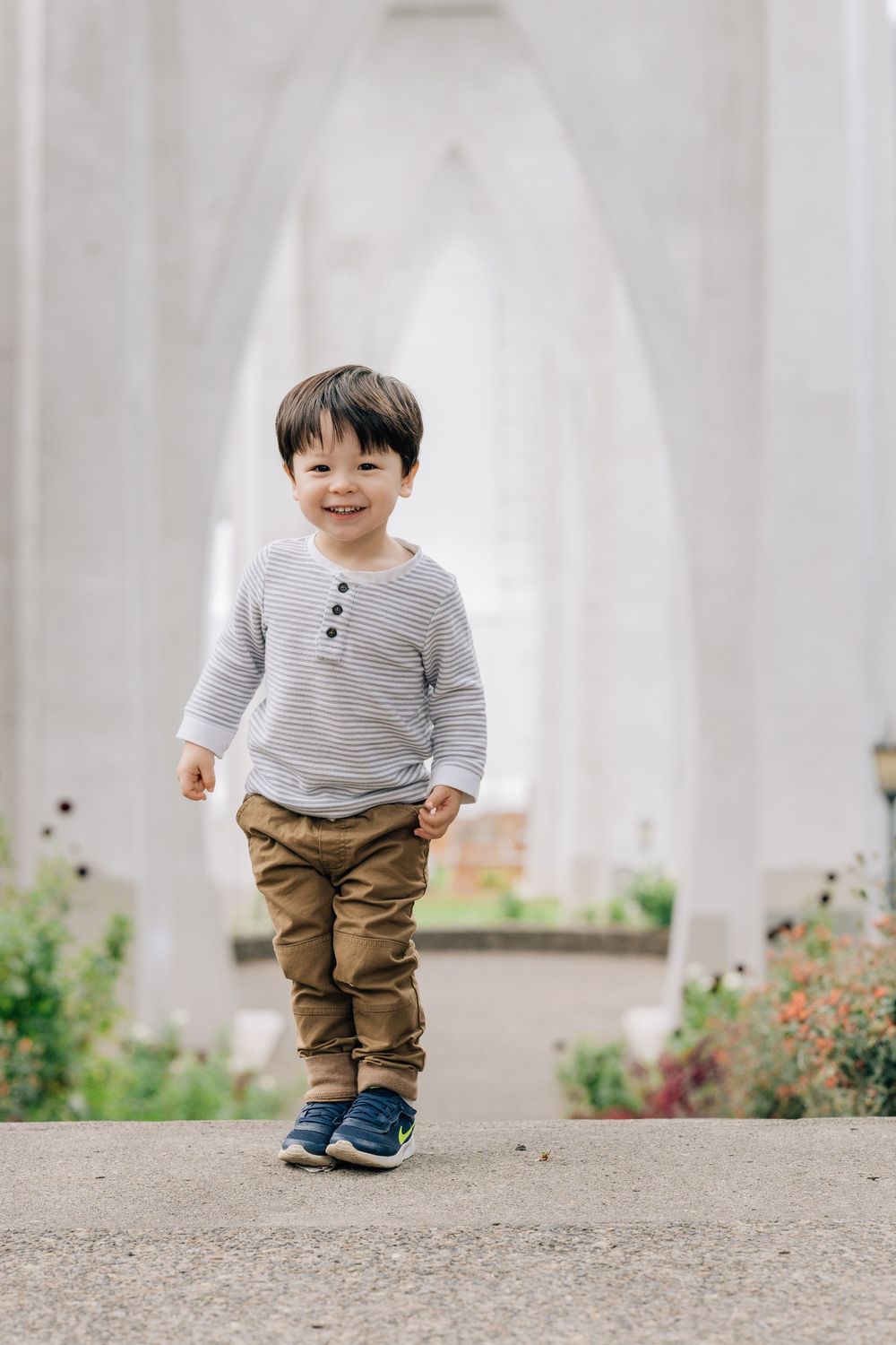 Happy toddler boy in striped shirt and khaki pants walking under white arched bridge outdoors.