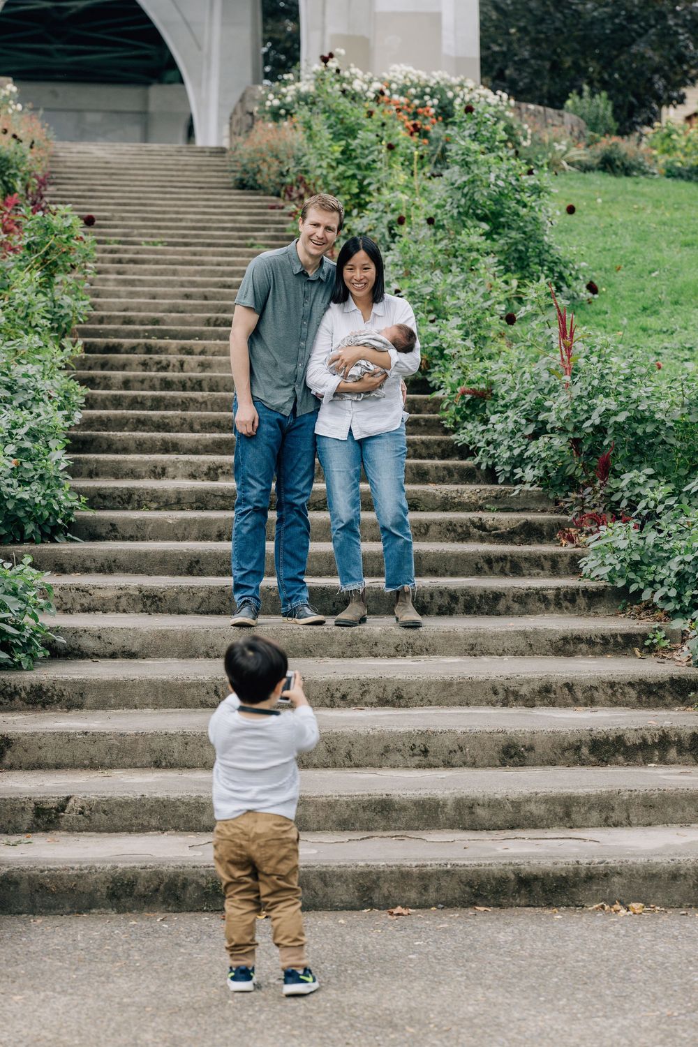A toddler photographs his parents standing on garden steps holding a newborn baby.