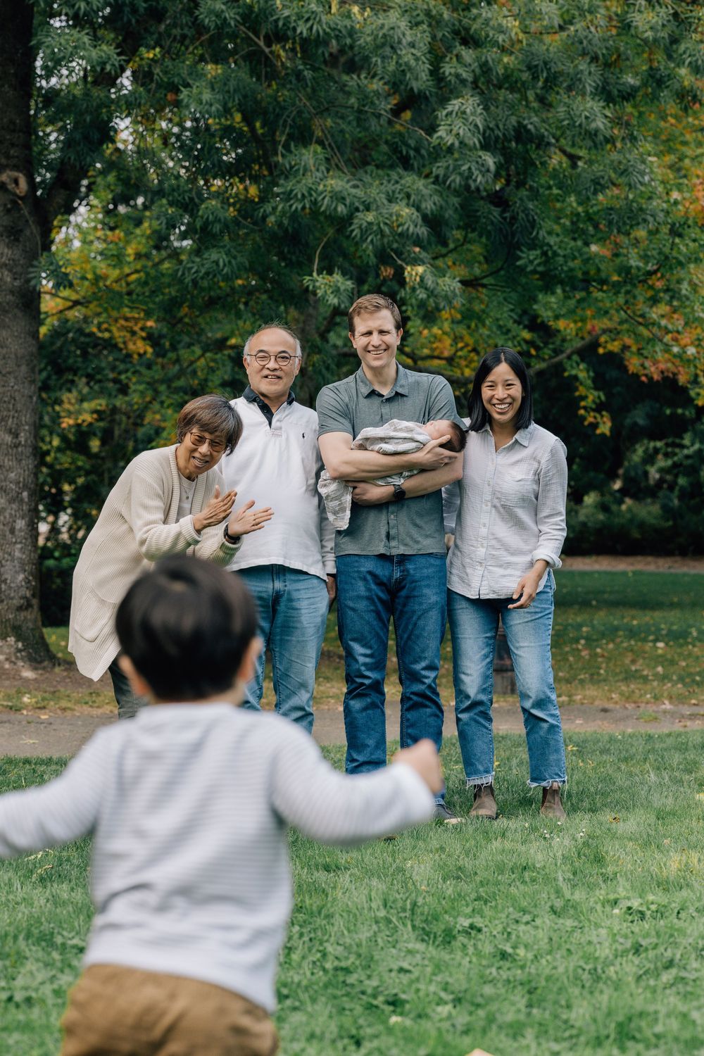 Multigenerational family smiles as toddler runs toward them in a lush green park.