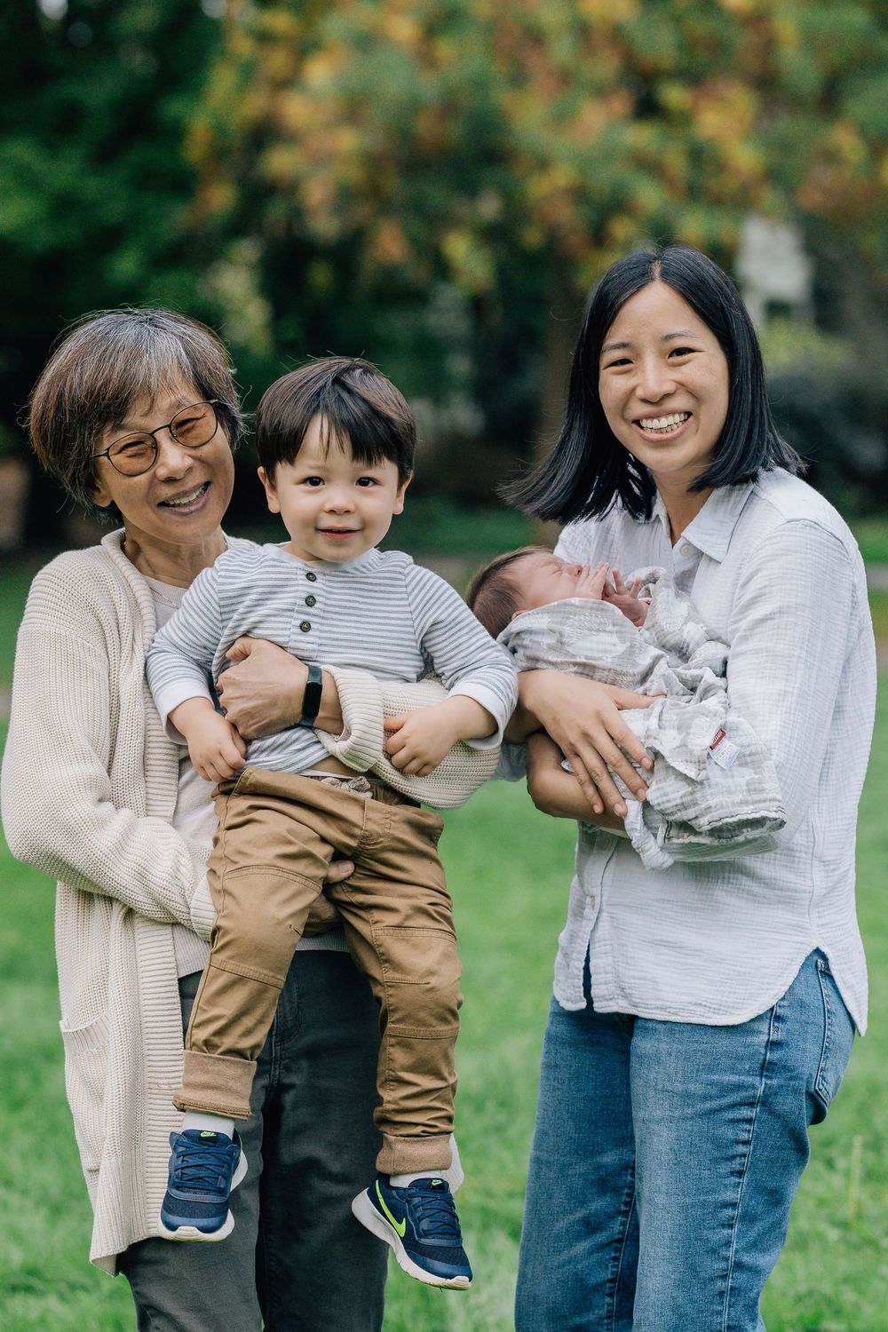 Grandmother holds toddler boy while mother cradles newborn baby outdoors in a park.