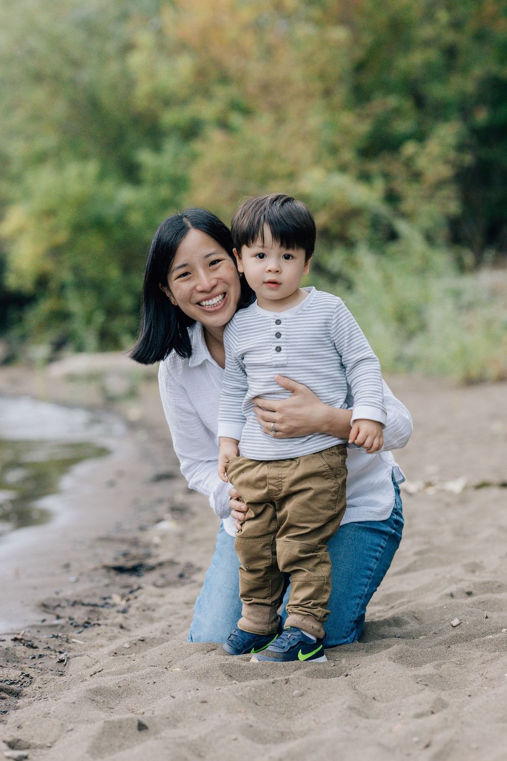 Smiling Asian mother kneels behind her toddler son on a sandy beach path.