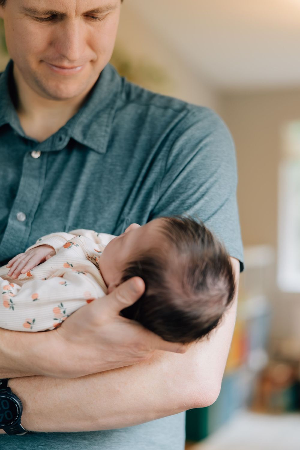 Father in gray polo shirt tenderly holds sleeping newborn baby in his arms.