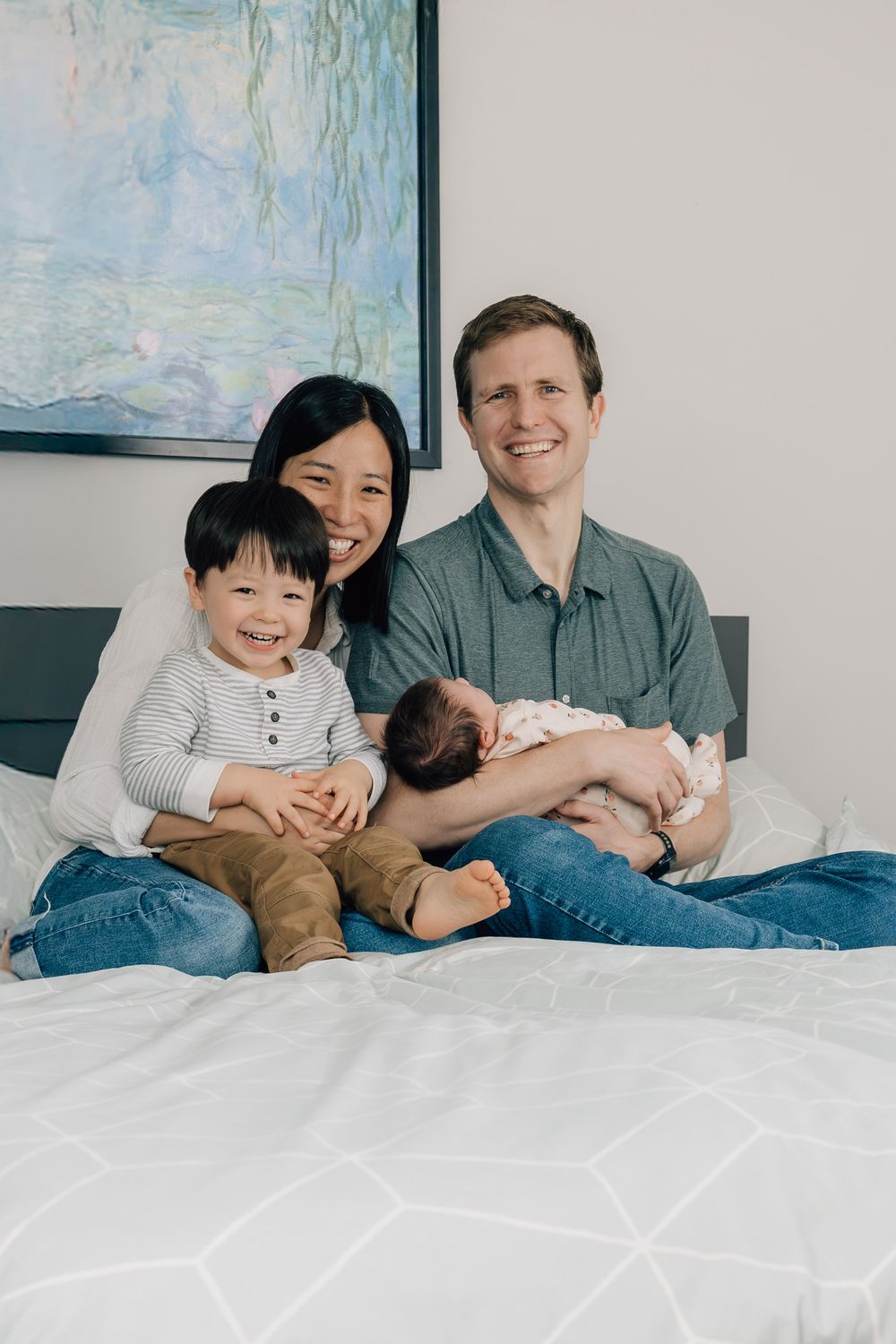 Happy family of four sits on a bed, parents smiling with toddler and newborn.