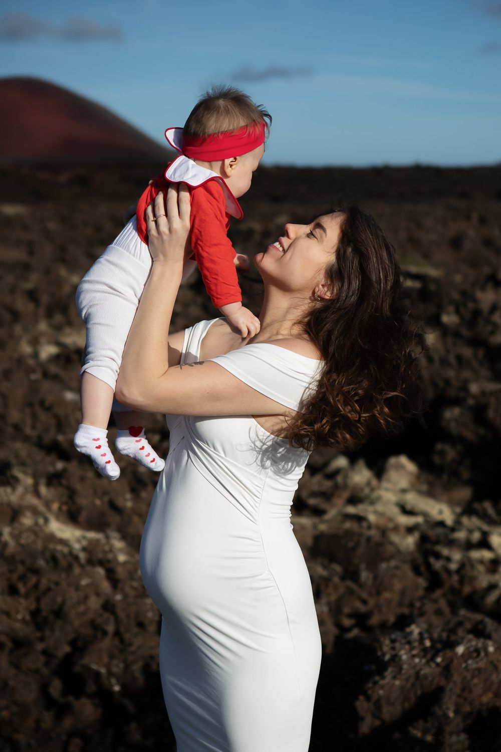 A pregnant woman in a white dress lifts a baby wearing red against a rocky landscape at sunset in Lanzarote
