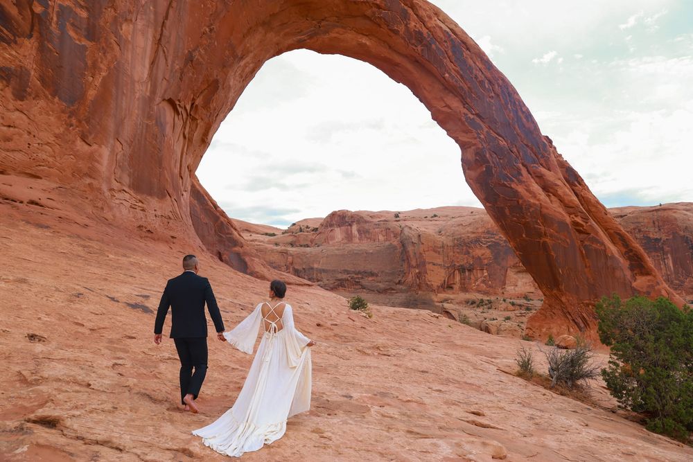 A couple in wedding attire walks beneath towering red rock formations in a desert landscape.