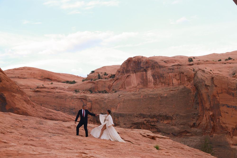 Wedding couple poses for photos among dramatic red rock formations in a desert landscape at sunset.