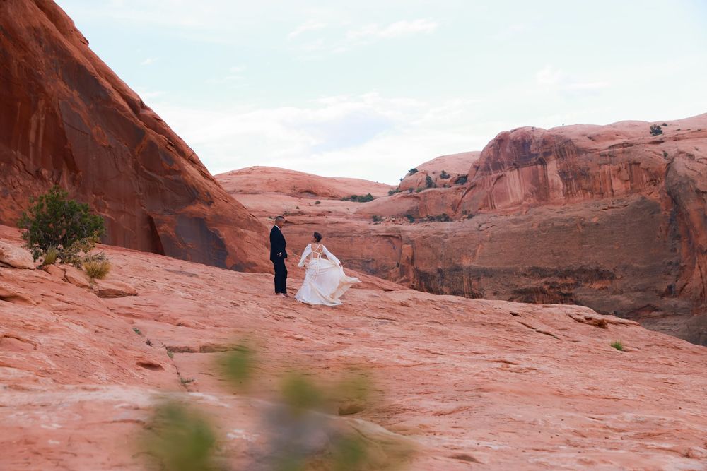 Two figures in formal attire stand together on red sandstone formations in a desert landscape.