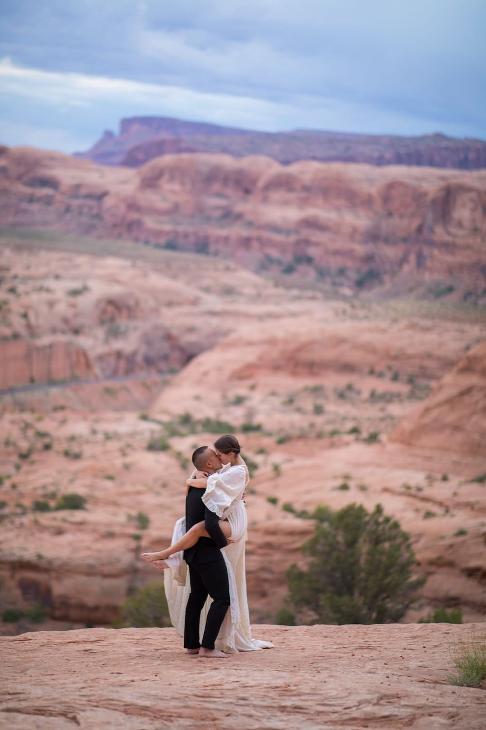 A couple embraces on red rock formations in the desert landscape of Utah's Arches National Park at sunset.