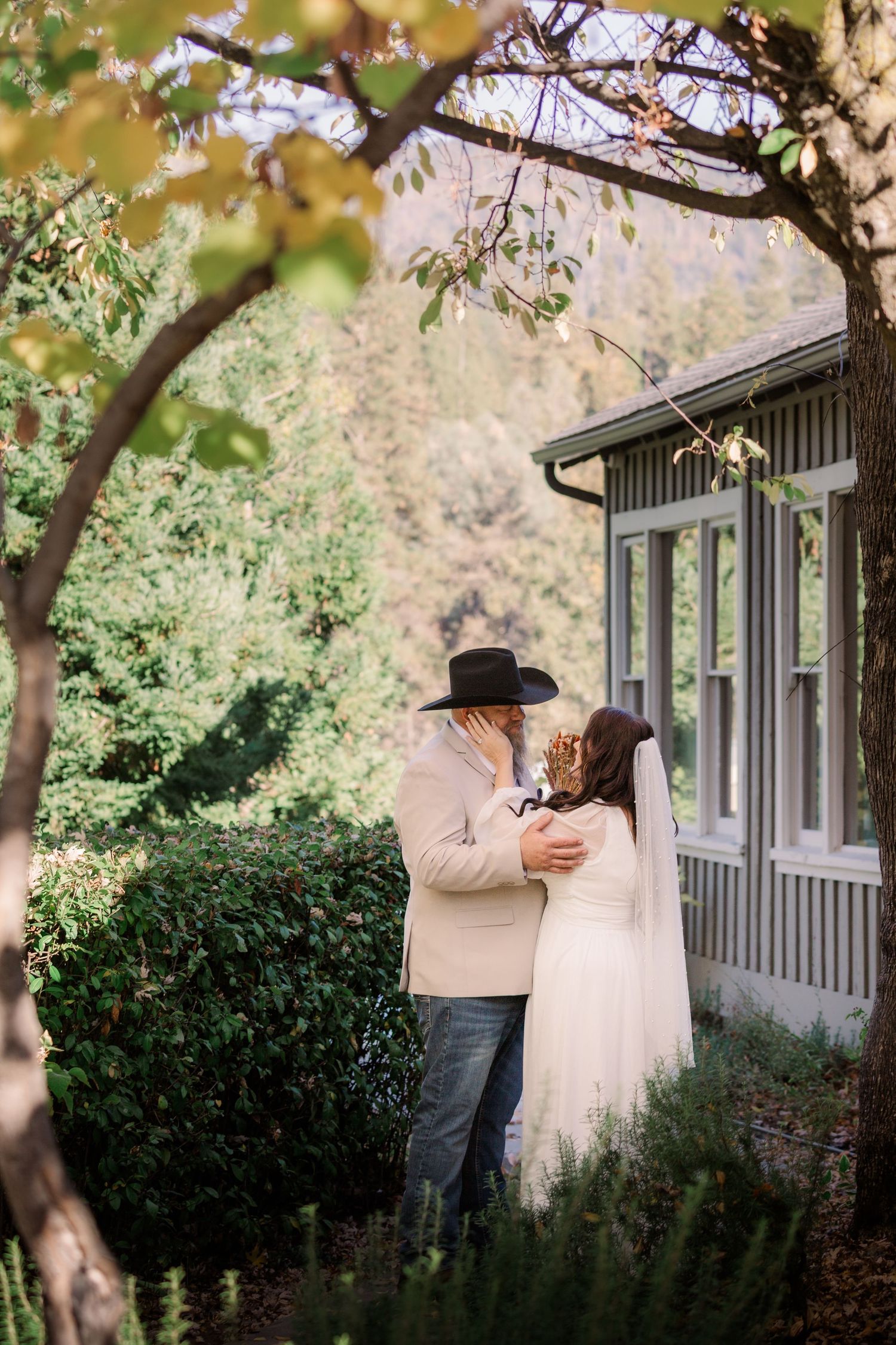 A couple shares an intimate moment in a garden setting with autumn foliage and a rustic building in the background.