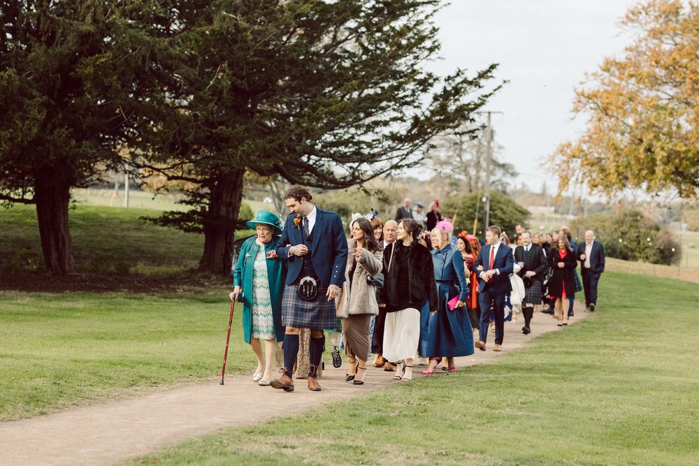 Wedding guests walk in a line along a grassy path at an outdoor venue during an autumn celebration.
