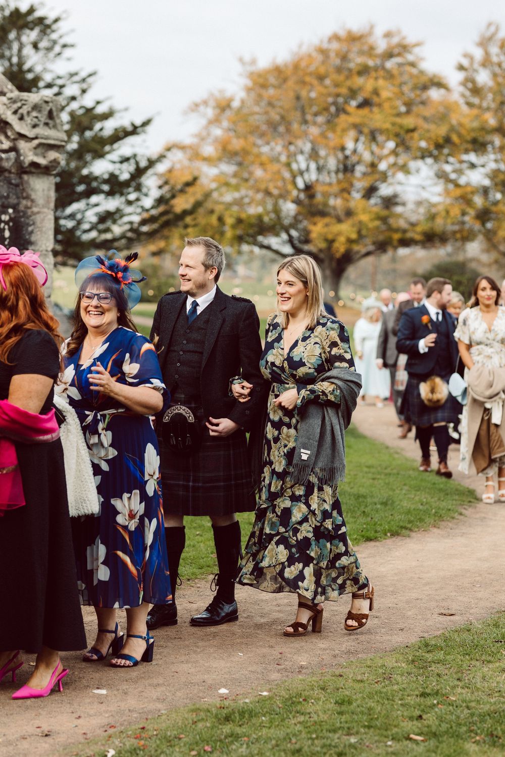 Wedding guests mingle and dance outdoors on a grassy path during an autumn celebration.