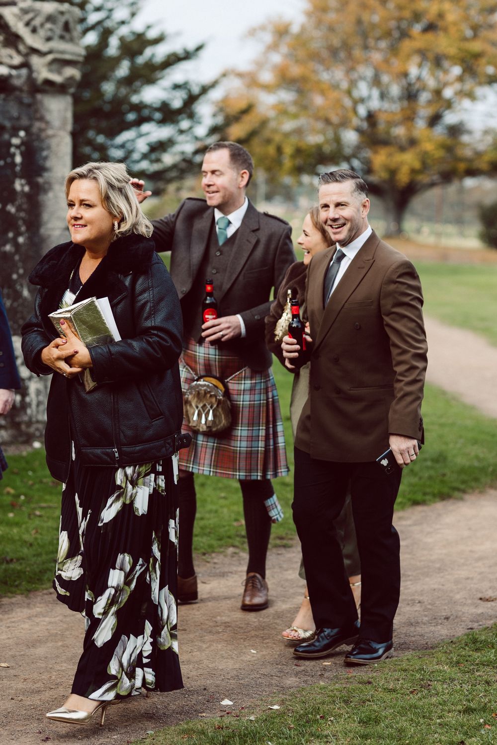 Wedding guests in formal attire walk along an autumn path with one wearing a traditional Scottish kilt.