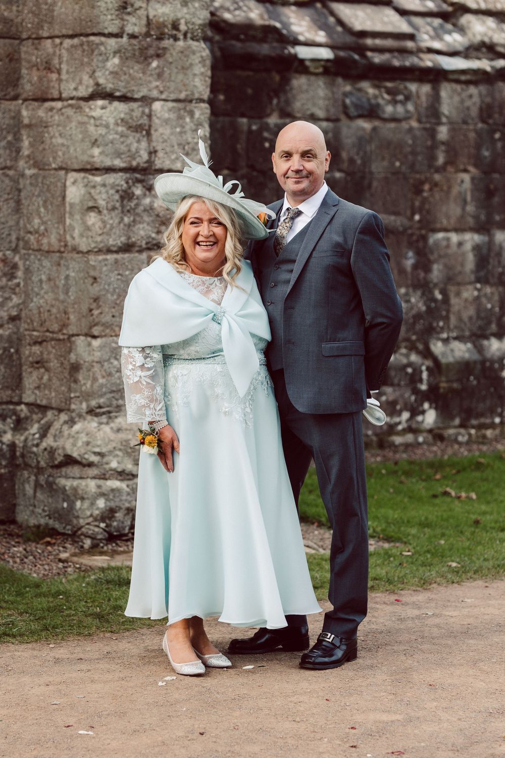 A happy couple in formal attire poses together outside against a stone wall in a series of candid wedding photos.