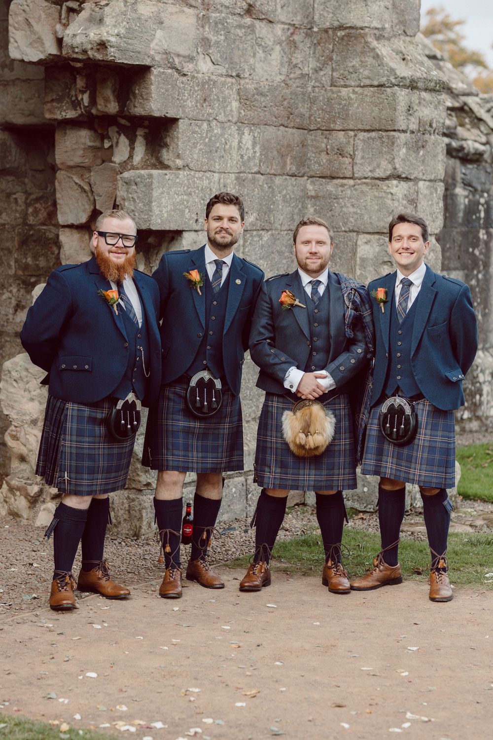 Groomsmen in matching navy suits and Scottish kilts stand against a stone wall backdrop.