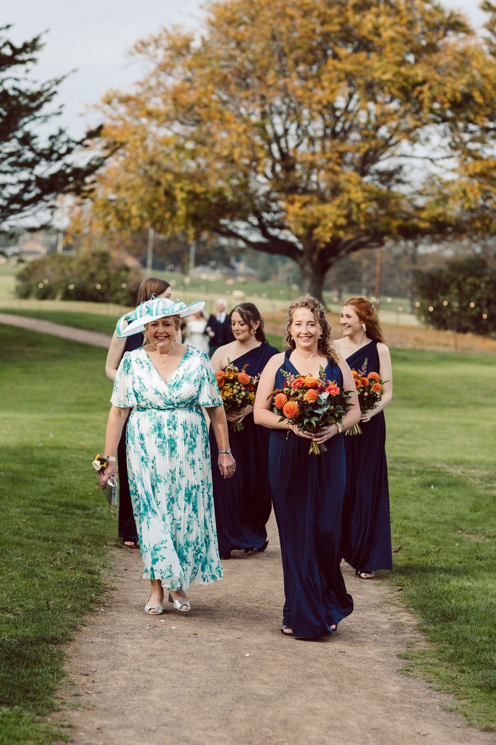 Bridesmaids in navy dresses carrying orange bouquets walk along an outdoor path in autumn.