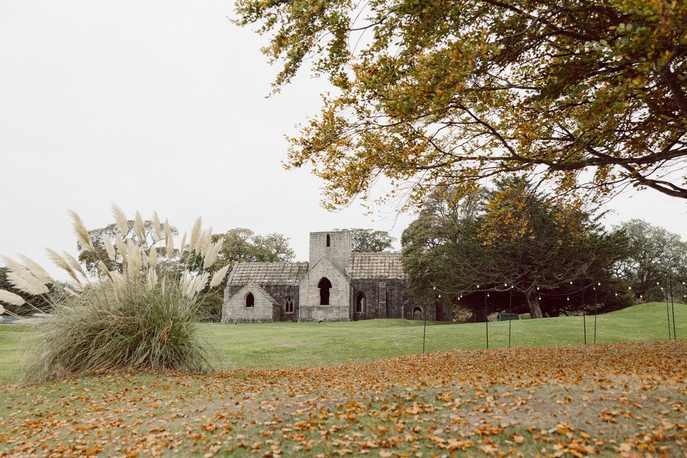 A rustic stone church building surrounded by autumn leaves and ornamental grasses in a peaceful countryside setting.
