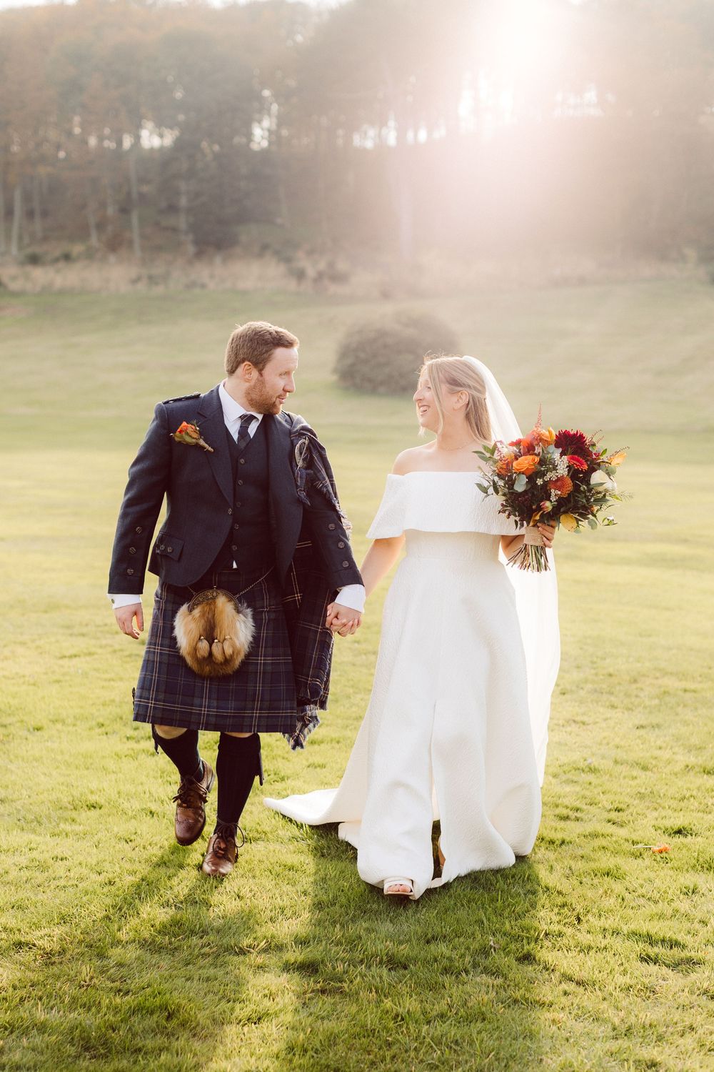 A couple in wedding attire walks through a sunlit meadow, with the bride's white dress flowing behind them.
