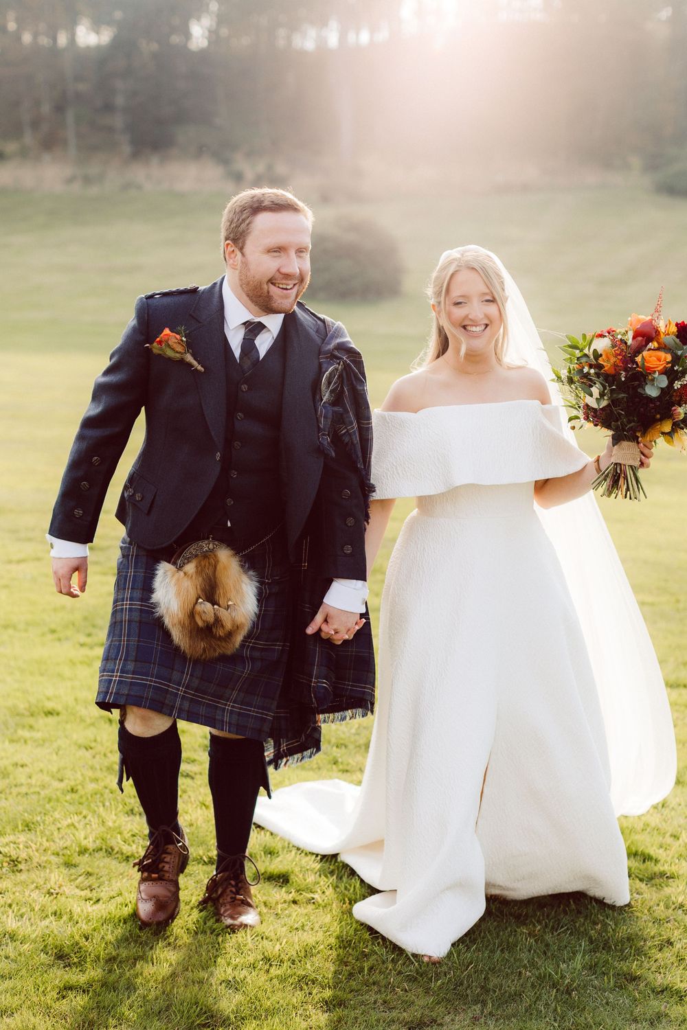 A couple in wedding attire walks together on a grassy lawn during their Scottish-themed outdoor celebration.