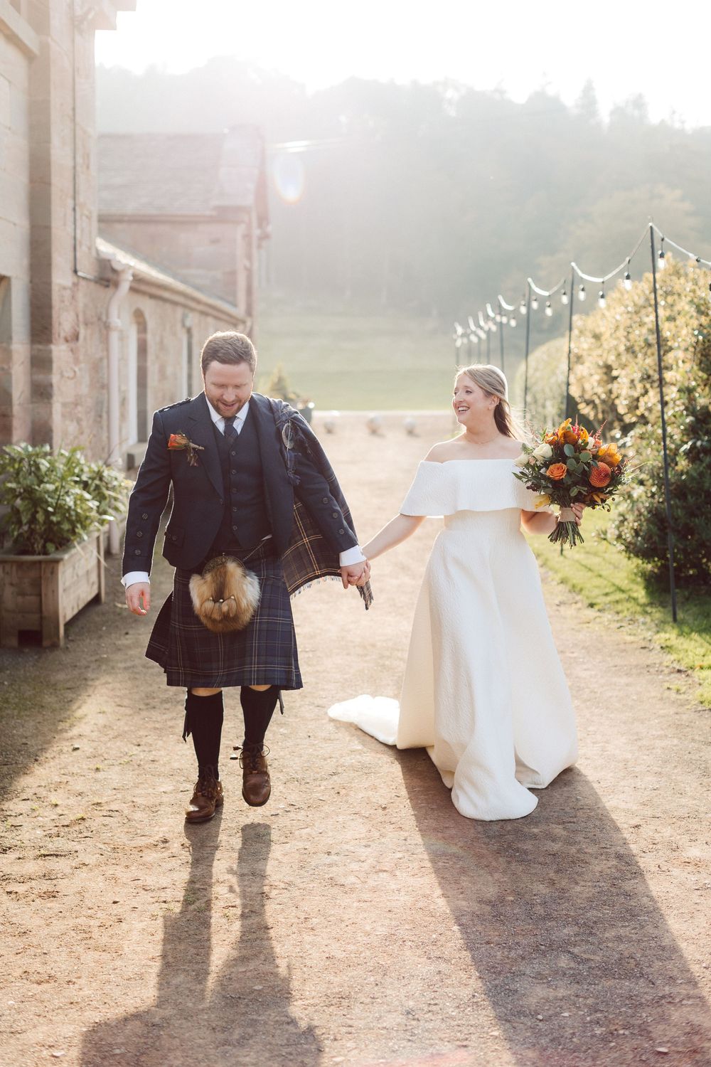 A couple in wedding attire walking and dancing together on a sunlit dirt path surrounded by greenery.