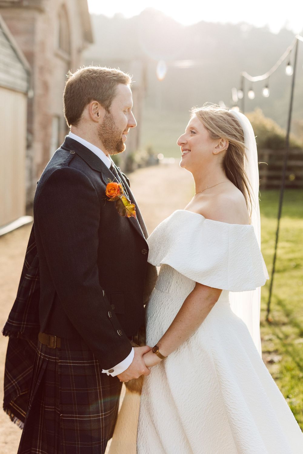 A romantic wedding couple shares joyful moments in golden sunlight while wearing traditional Scottish attire and white gown.