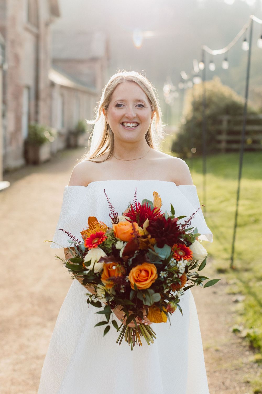 Beautiful autumn wedding bouquet with orange roses, burgundy dahlias and wildflowers held against white off-shoulder dress.