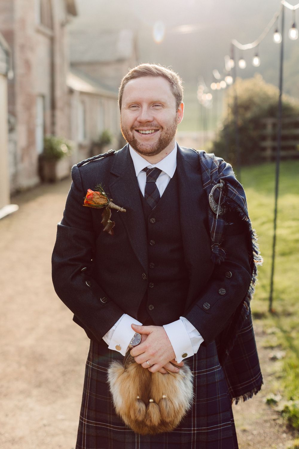 A groom in a traditional Scottish kilt and formal black jacket holds a fluffy ginger cat during an outdoor photoshoot.