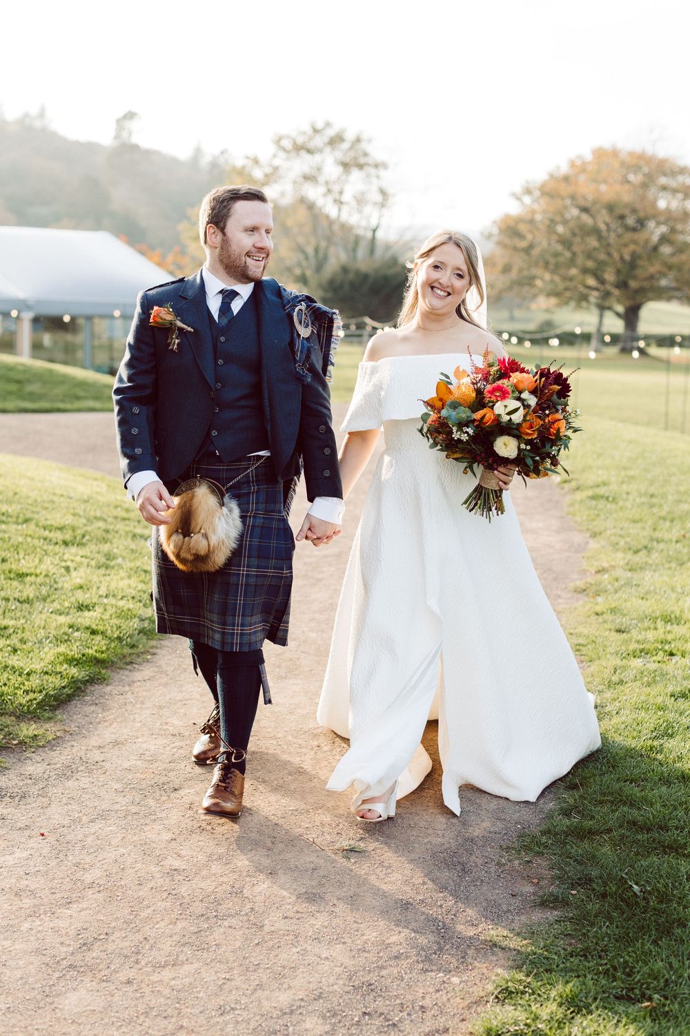 A couple walks hand in hand along a scenic countryside path during their autumn wedding celebration.