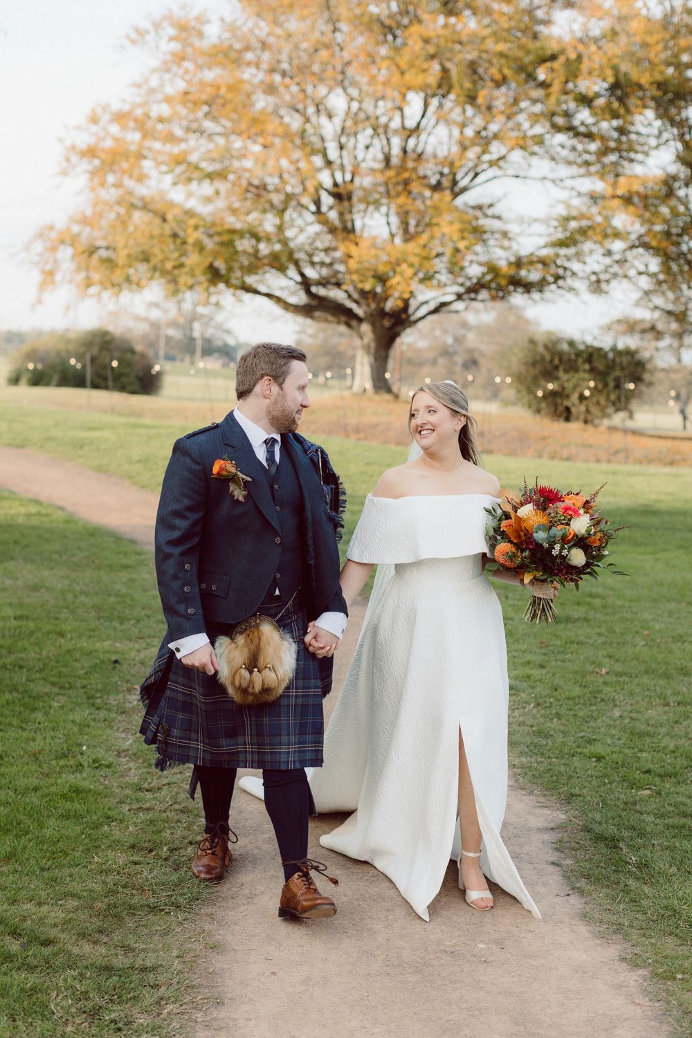 A couple in wedding attire walk together on a grassy hillside during autumn with golden trees in the background.