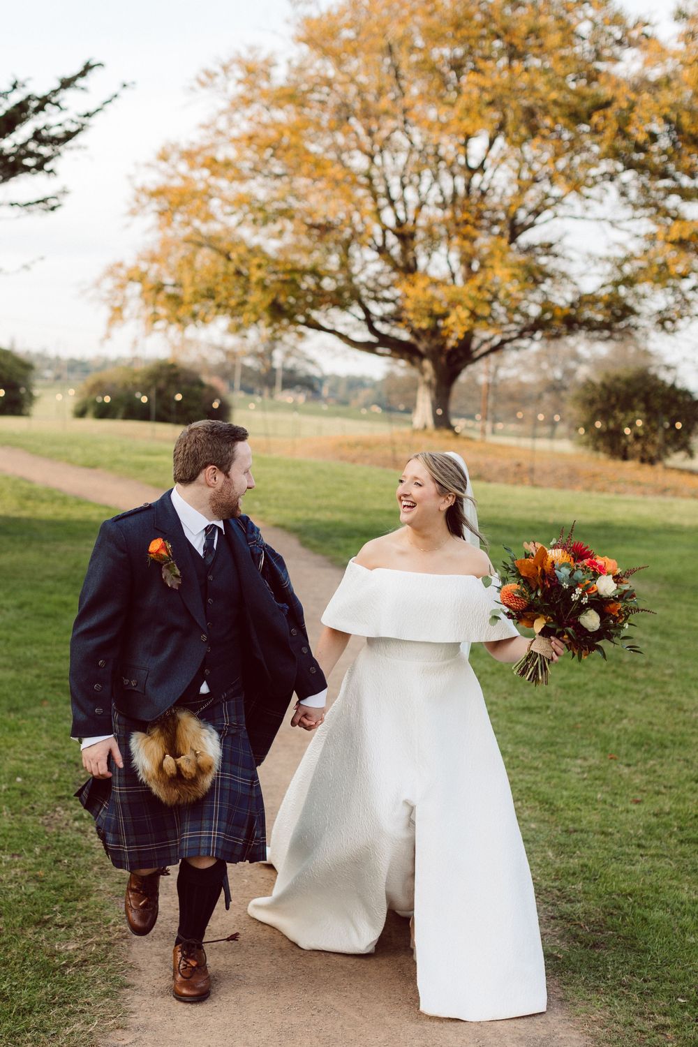 A couple in wedding attire sits together on a bench under autumn trees with orange and yellow foliage.
