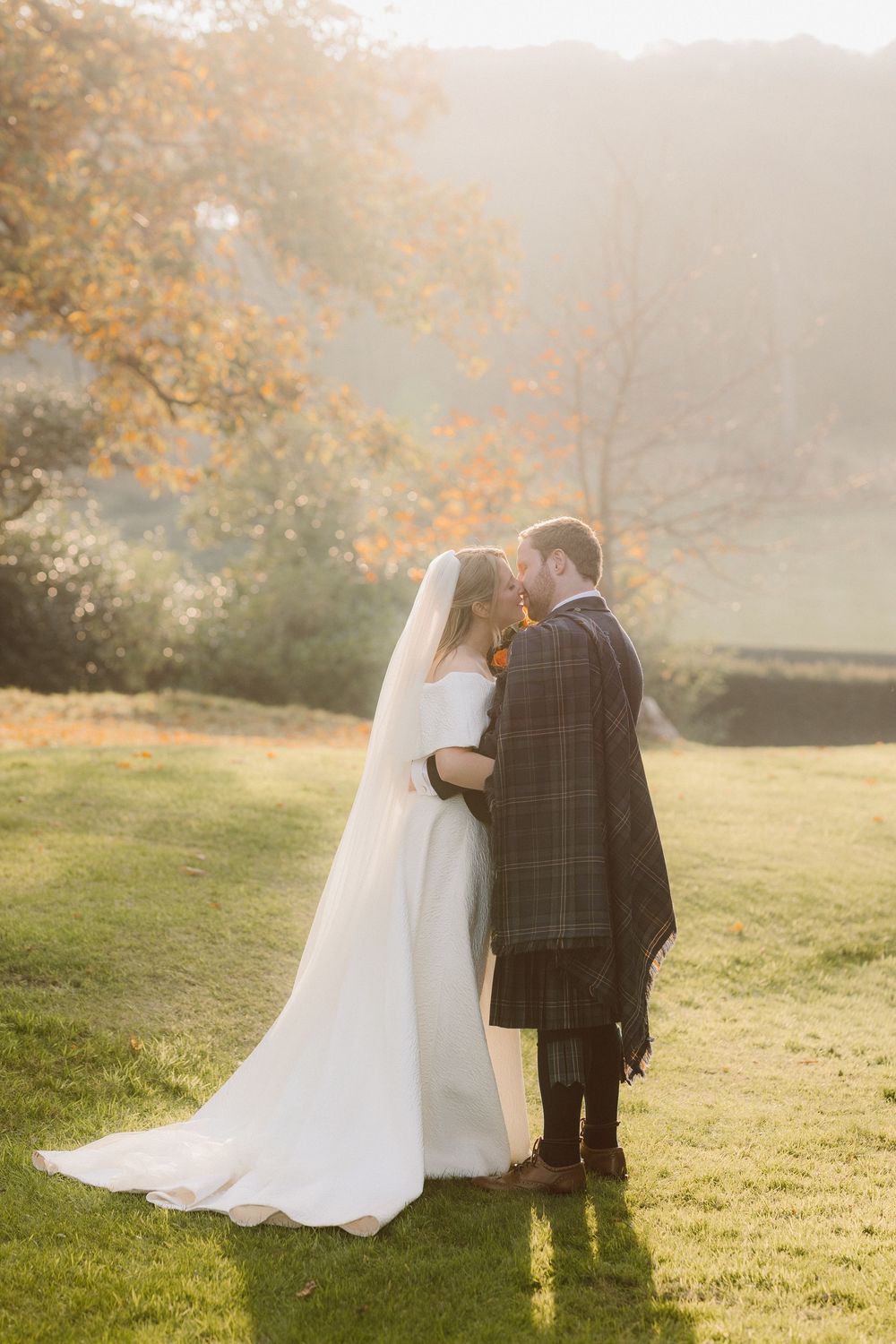 Newlyweds sit together on grassy hillside during golden sunset with autumn trees in background.