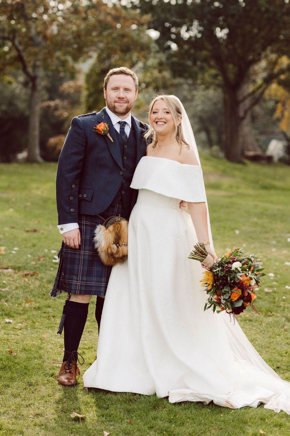 Wedding couple poses outdoors in autumn, with bride in white dress and groom wearing traditional Scottish kilt.