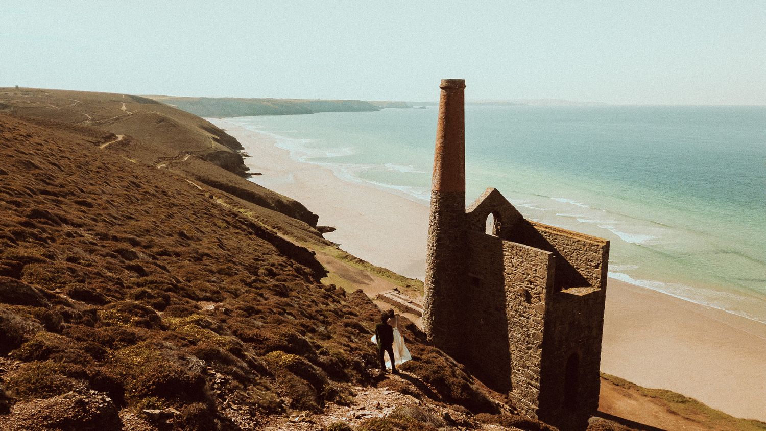 Historic mining ruins stand atop rugged coastal cliffs overlooking a sandy beach and turquoise ocean.