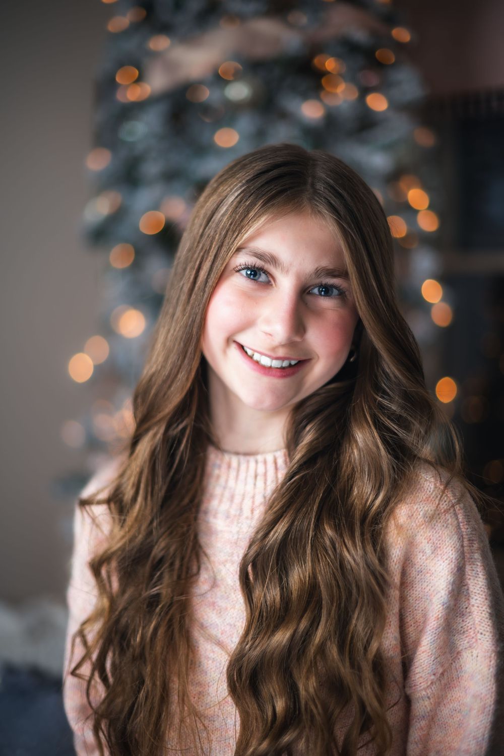 Portrait against blurred holiday lights wearing a cream sweater with long wavy brown hair.