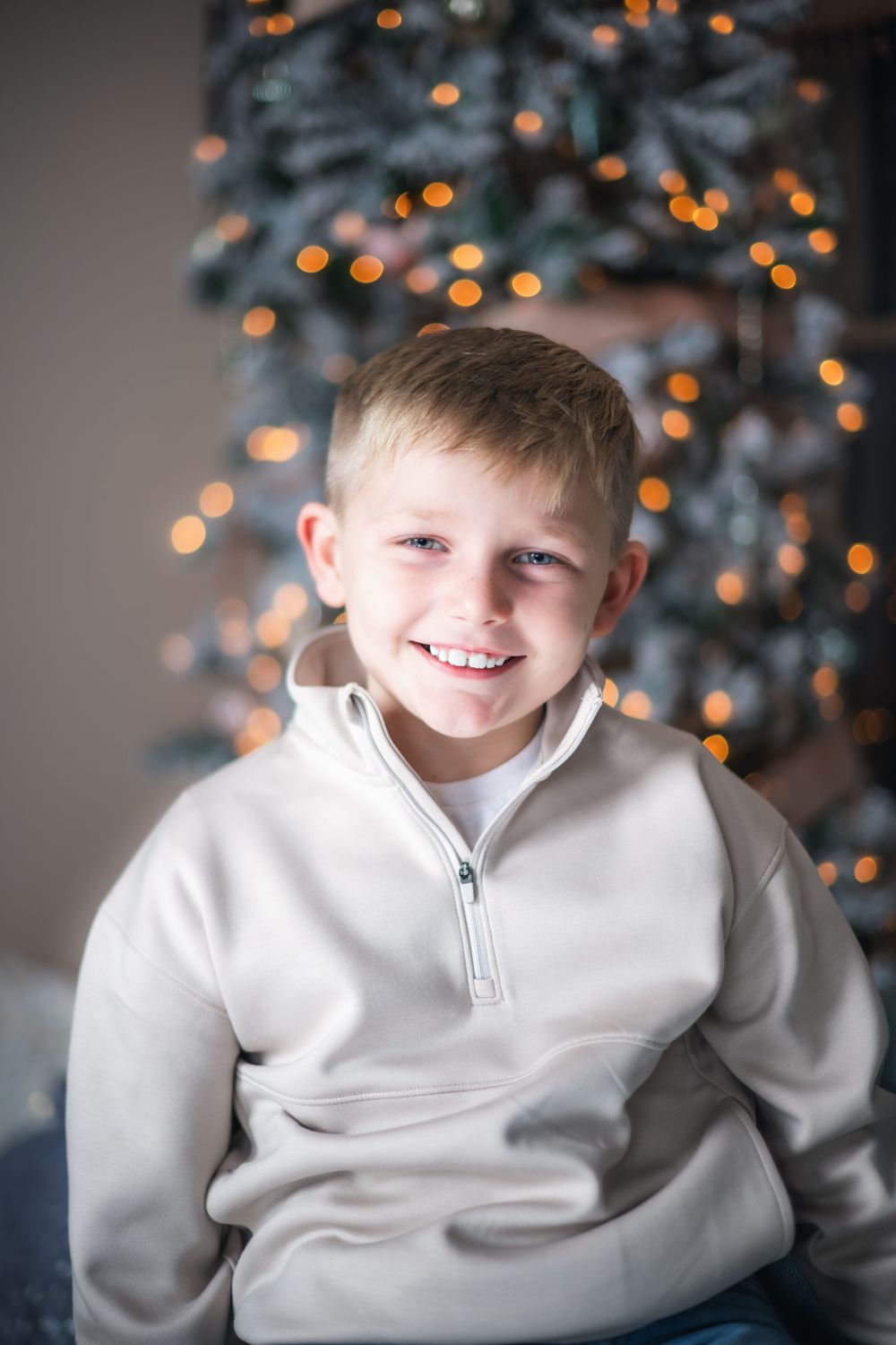 A child wearing a light gray quarter-zip sweater poses in front of a Christmas tree with twinkling bokeh lights.